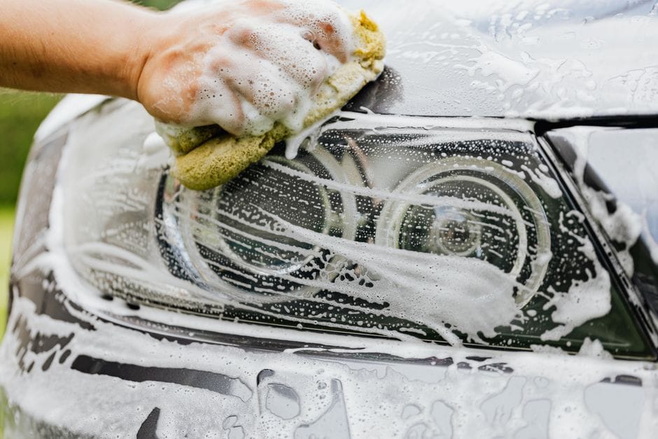 Hand scrubbing a car headlight with sponge and soap suds, detailing process.